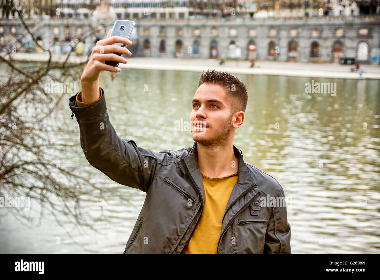 Tre quarti di lunghezza di luce dai capelli marrone giovane uomo indossando giacca grigia e jeans denim tenendo selfie foto con il suo smartphone Foto Stock