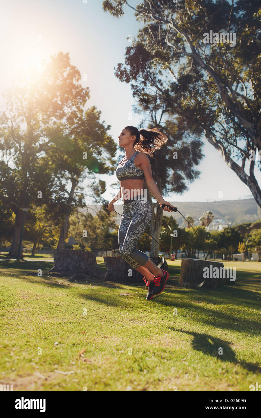 Lunghezza piena ripresa esterna della giovane donna saltando con un salto con la corda all'esterno. Donna facendo un allenamento fitness in un parco su una soleggiata da Foto Stock