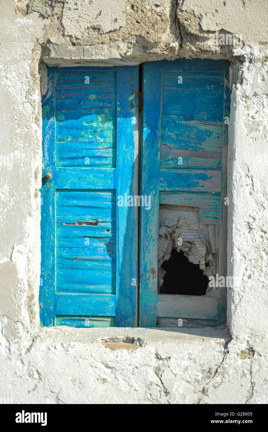 Un vecchio danneggiato finestra greco in stato di abbandono con stacca la vernice blu sul clappe chiuse in un bianco parete rappresentata in Grecia Foto Stock