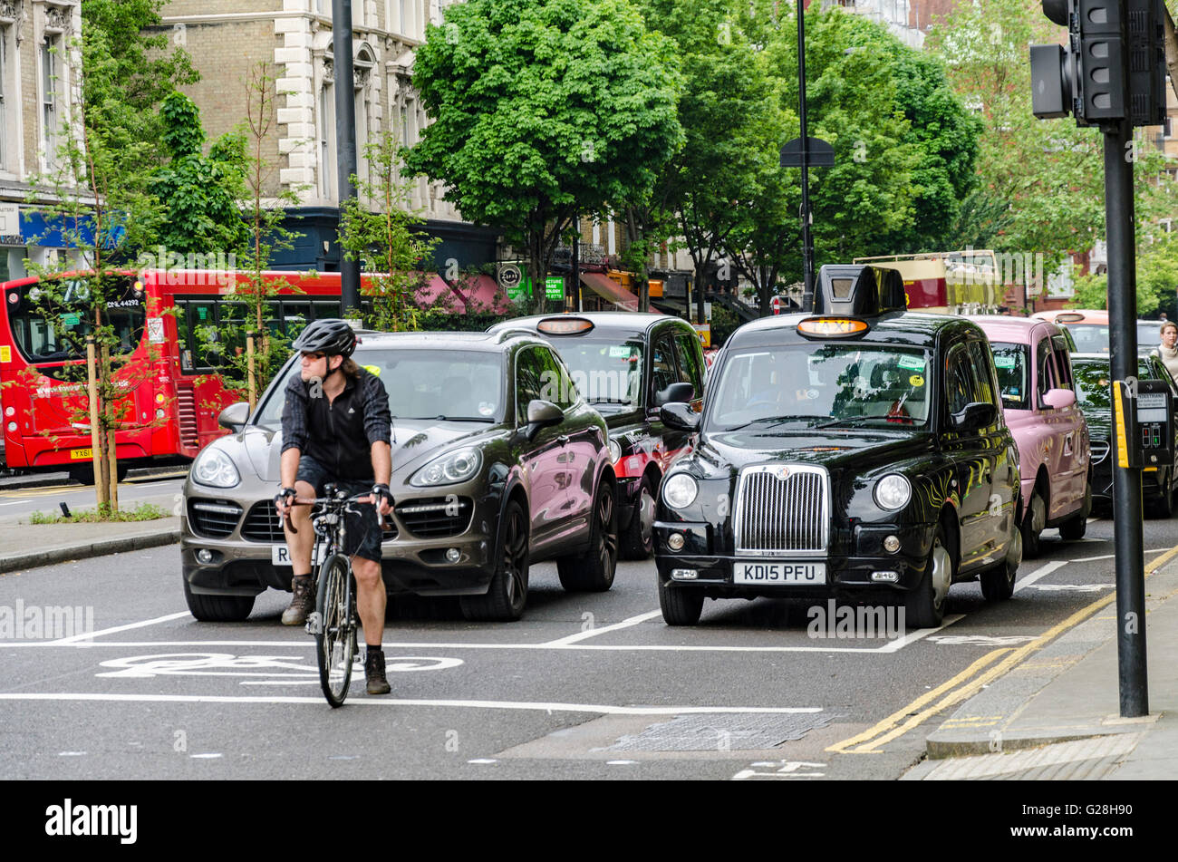 Un ciclista attende alla testa di una coda a un semaforo rosso a Londra davanti a vetture e taxi. Foto Stock