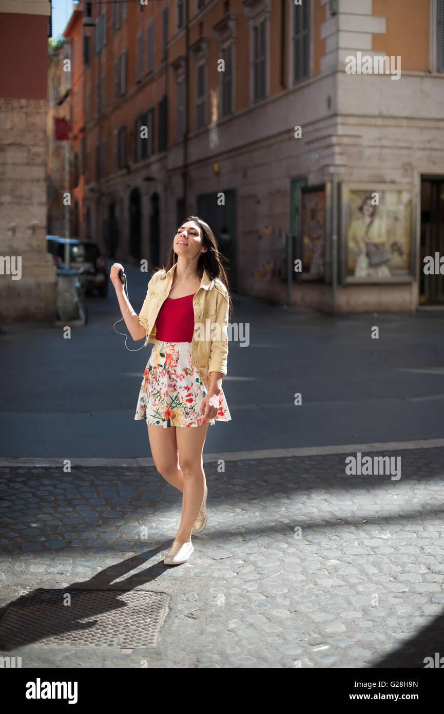 Sorridente giovane donna allegra ascoltando musica con le cuffie mentre si cammina in una strada di città Foto Stock