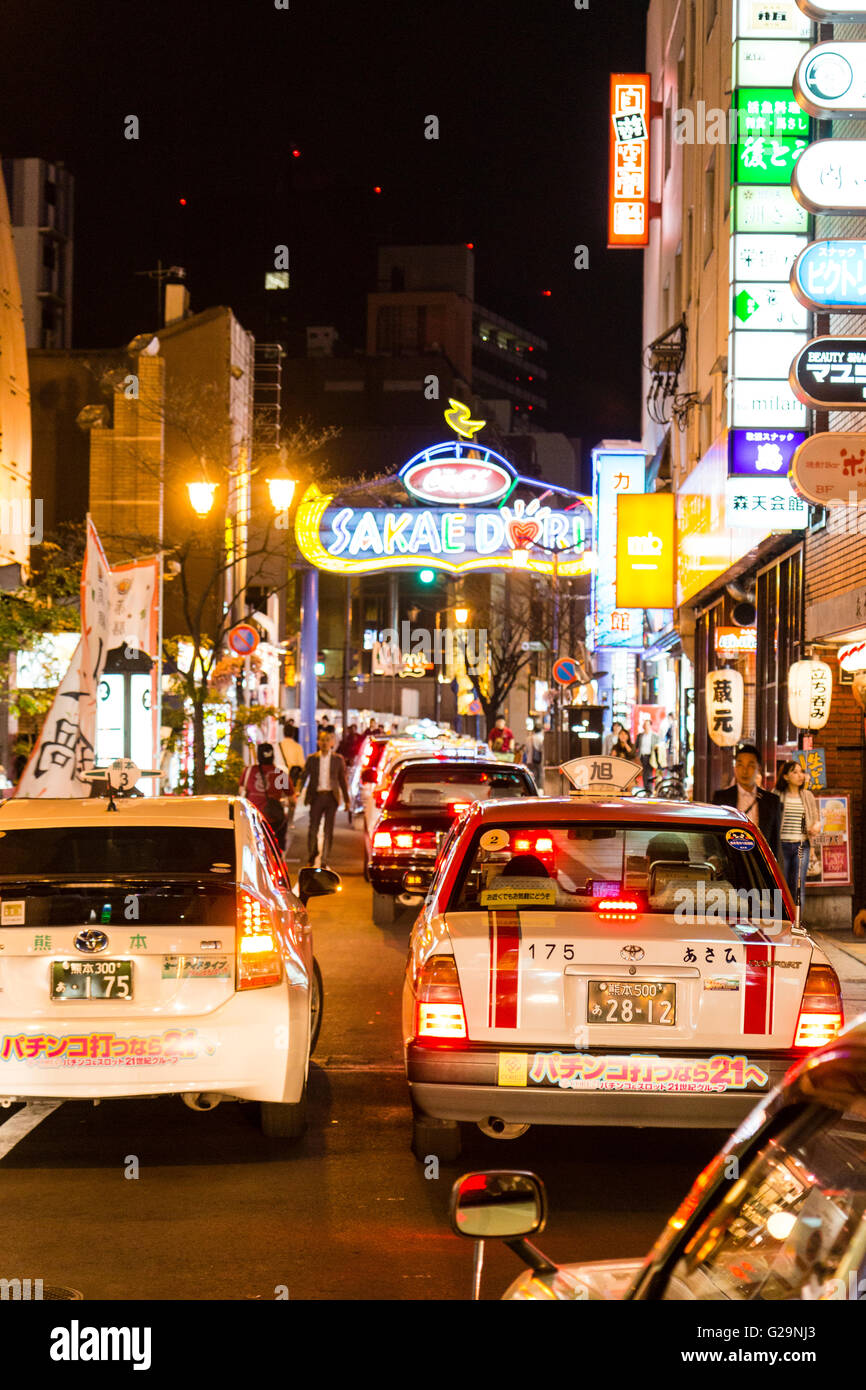 Giappone Kumamoto, Centro citta'. Vista lungo la stretta strada congestionata con la linea di auto e taxi-cabine di notte tempo. Molte insegne su edifici. Foto Stock