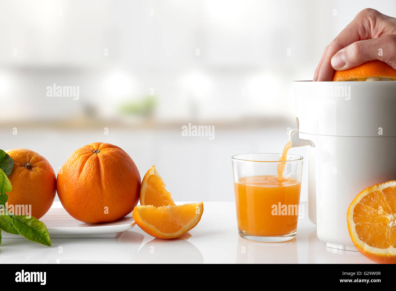Spremitura a mano un arancio con un spremiagrumi elettrico in un vetro in una cucina bianca tabella. Arance su una piastra e taglio. Posizione orizzontale Foto Stock
