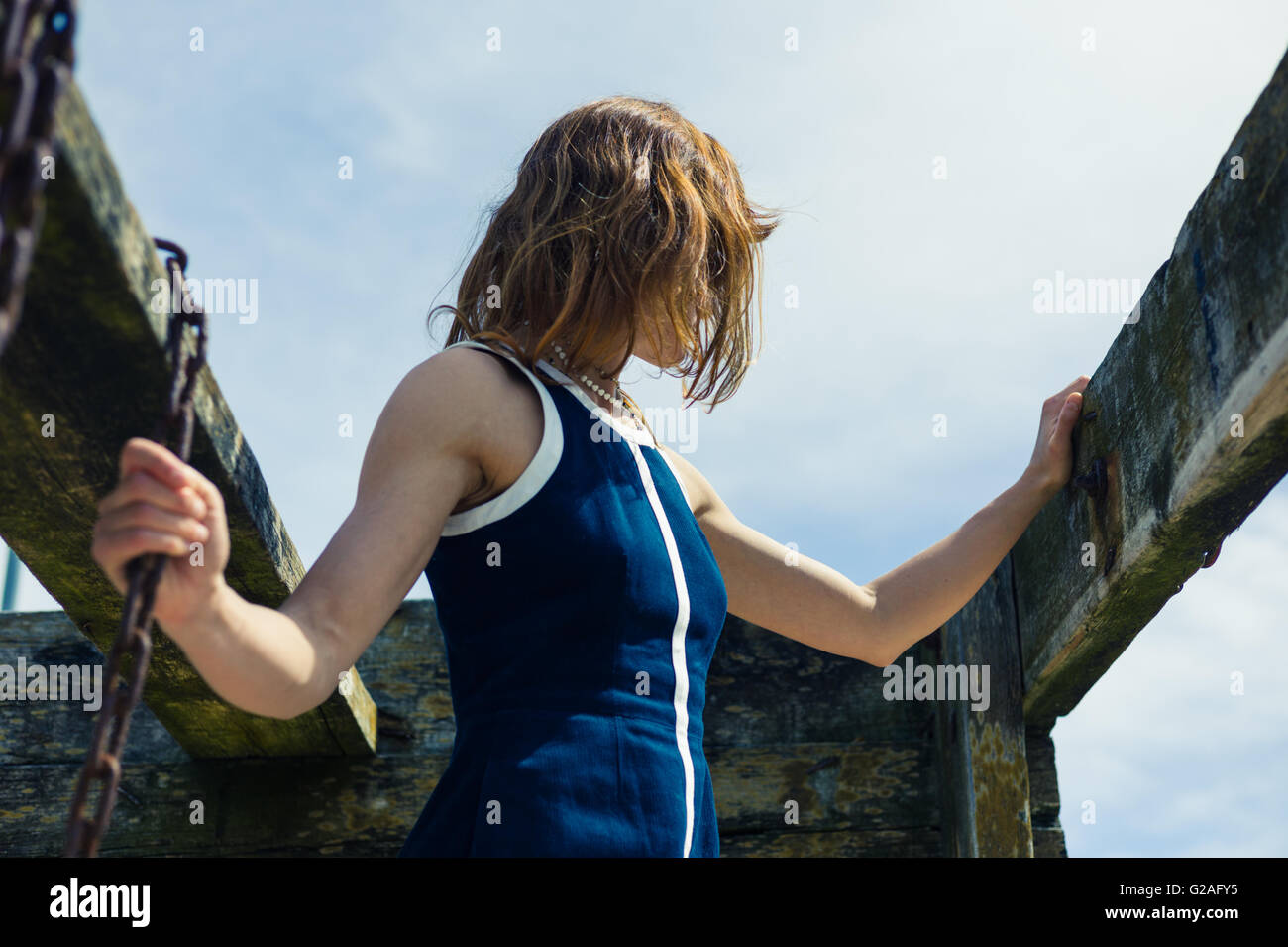 Una giovane donna che indossa un vestito blu è in piedi fuori in una giornata di sole da due travi di legno con rusty catene appese da loro Foto Stock