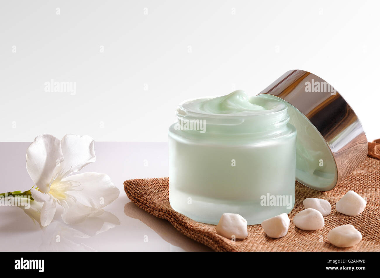 Vetro vaso aperto con viso o corpo crema sulla tela ruvida. con coperchio, pietre e fiori. Bianco sfondo isolato. Vista frontale. Foto Stock