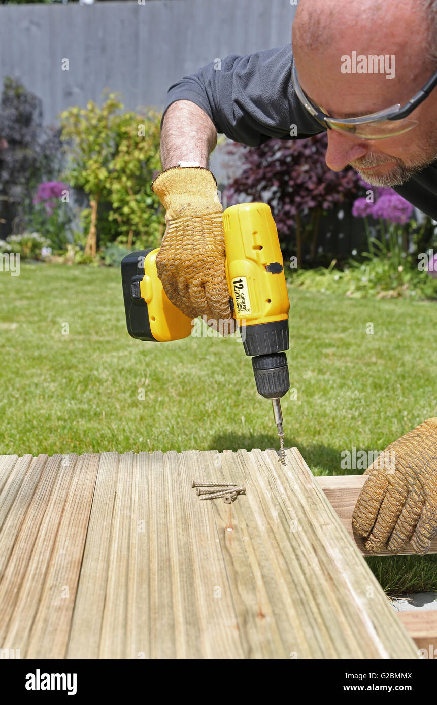 Un operaio utilizza un trapano senza filo per assemblare giardino decking in un giardino di Londra con un prato e arbusti in background Foto Stock