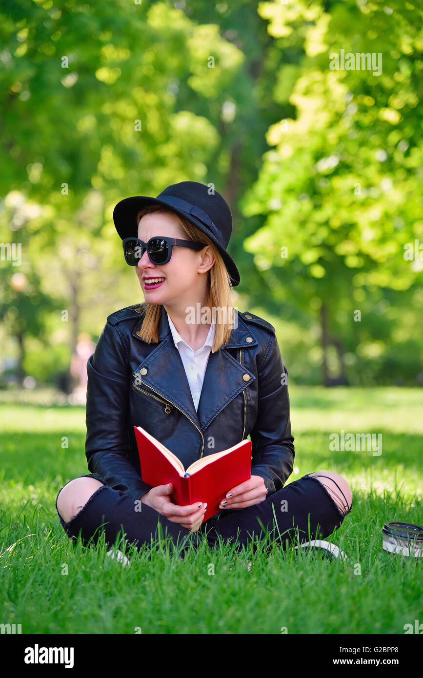 Hipster ragazza in hat seduto sull'erba con libro Foto Stock