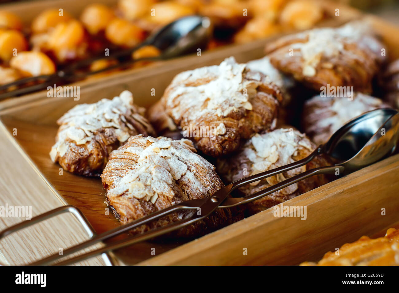 Croissant cosparso di mandorle e zucchero in polvere in un vassoio di legno con pinze in metallo Foto Stock