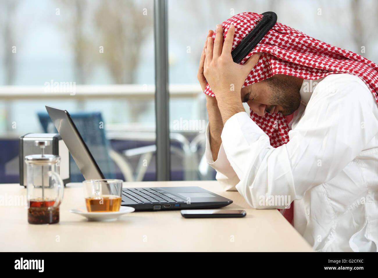 Profilo di una disperata e sola arab arabia uomo con un laptop online in una caffetteria con una finestra in background Foto Stock