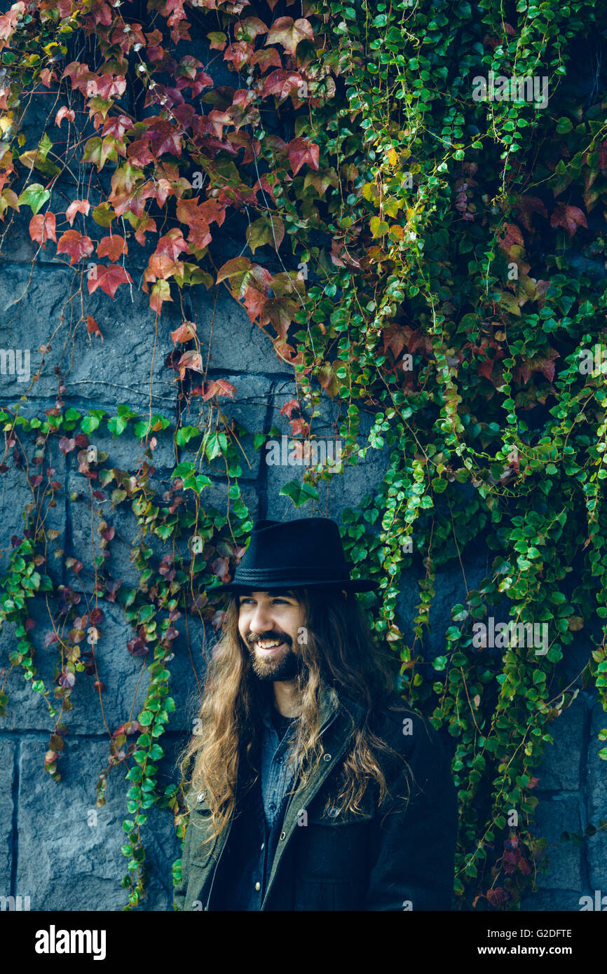 Ritratto di uomo sorridente con la barba e capelli lunghi in piedi contro la parete Vine-Covered Foto Stock