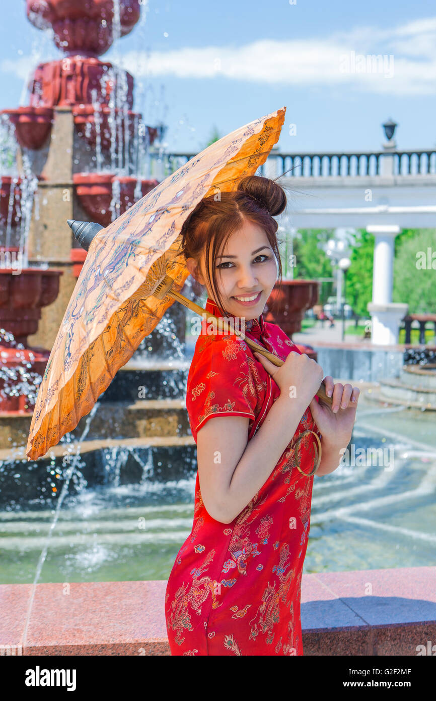 Bella ragazza asiatica in cinese tradizionale abito rosso con ombrello di bambù vicino alla fontana Foto Stock