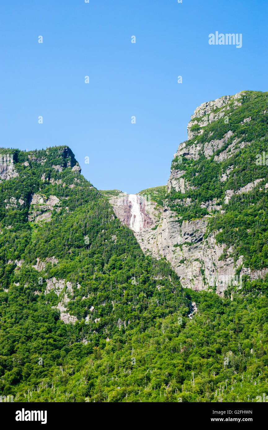 Ripide scogliere coperte di vegetazione con cascata in distanza che scorre dalla cima della montagna sotto il cielo blu chiaro. Foto Stock