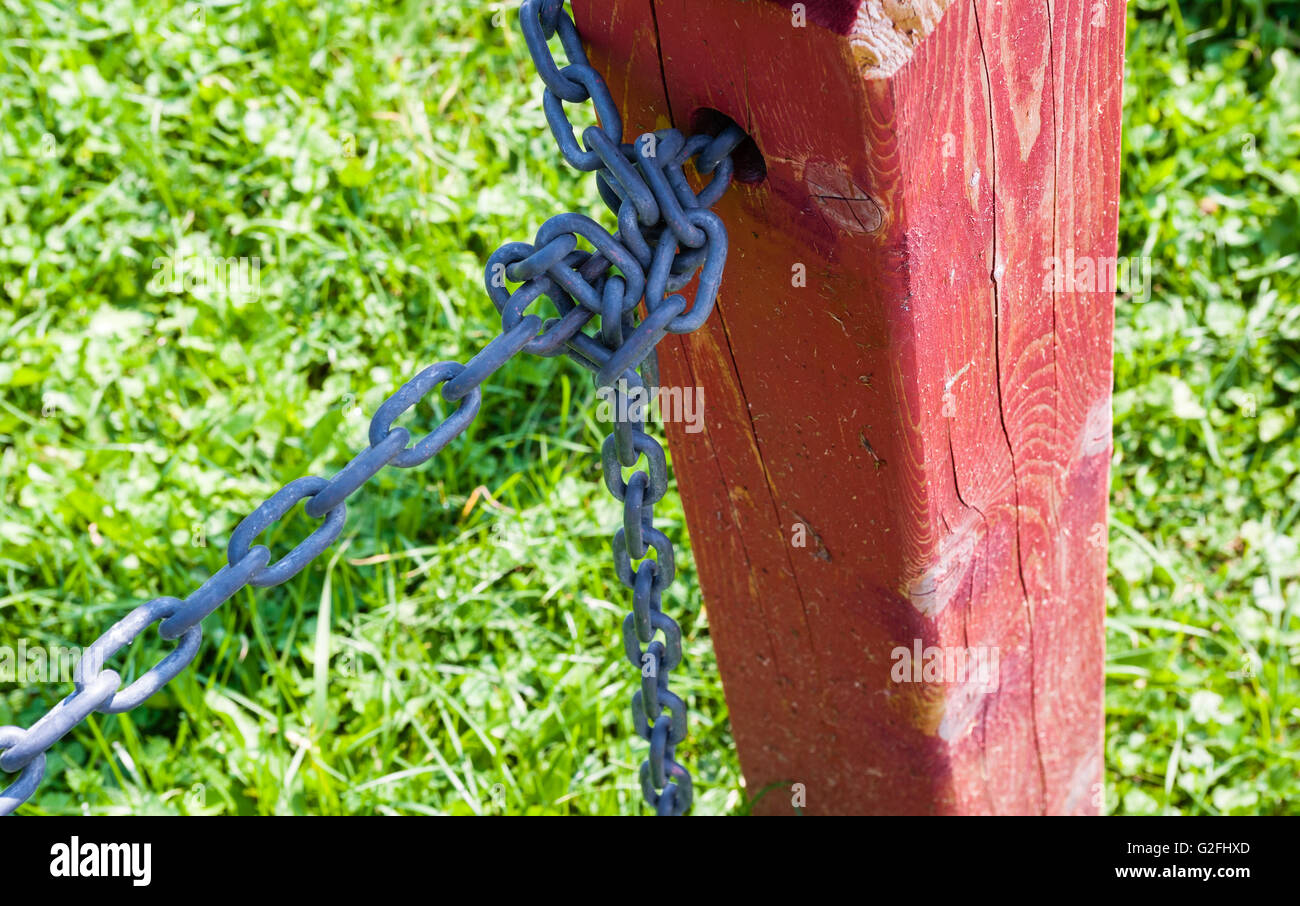 Dettaglio del recinto rosso post con catena metallica avvolta attraverso il foro nel post e legato, contro l'erba verde. Foto Stock