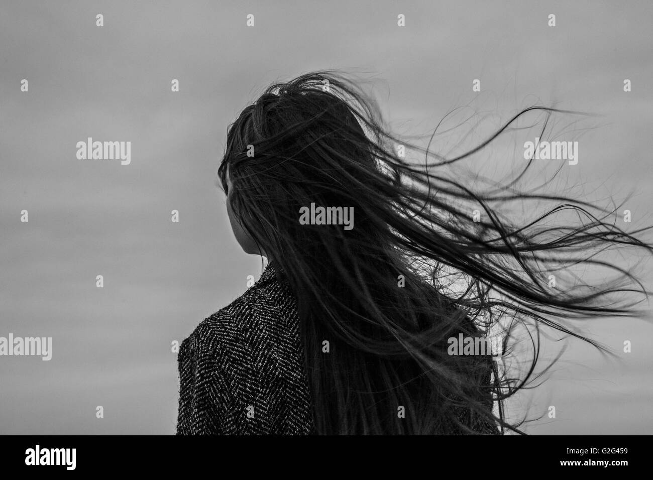 Giovane donna con capelli soffiando nel vento sulla giornata grigia, vista posteriore Foto Stock