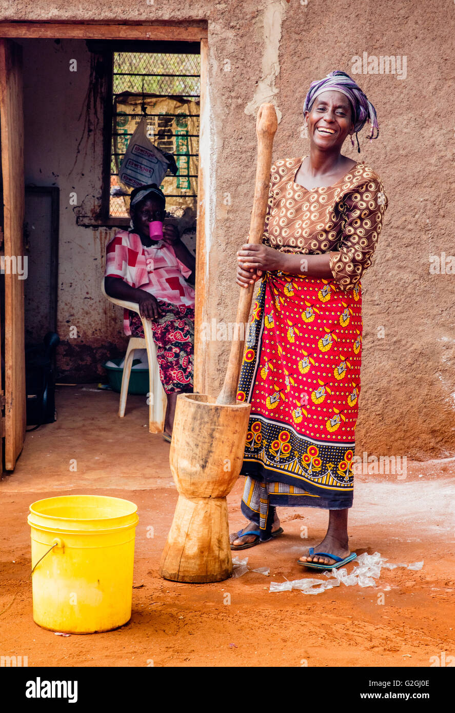 Allegro sorridente signora in una scuola in Kenya la preparazione di spezie con un gigante di pestello e mortaio mentre il suo collega le bevande di tè - Voi Foto Stock