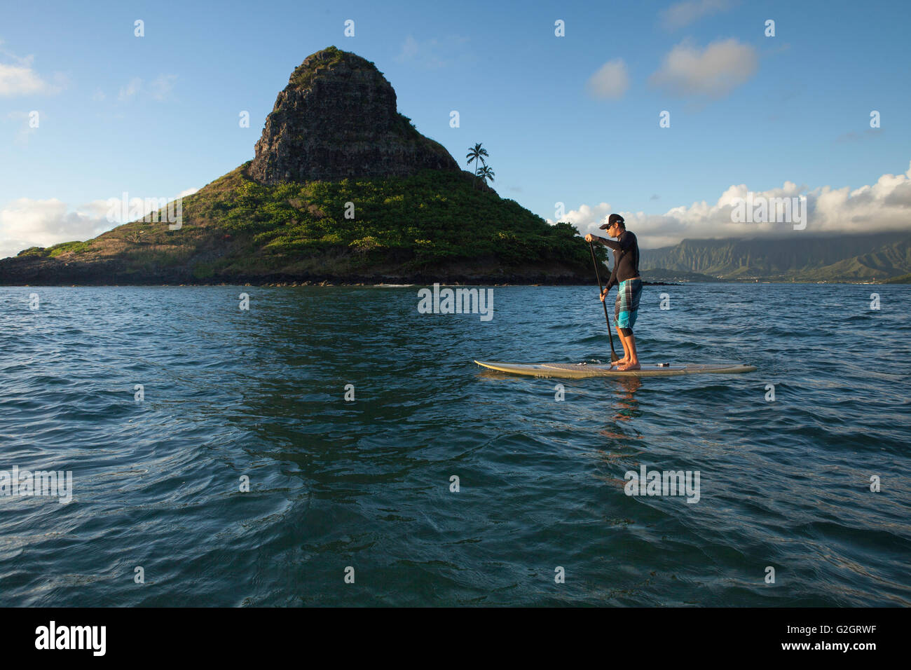 Sali a paddle boarding nella baia di Kaneohe, vicino a Kualoa e all'isola di Mokoli'i (precedentemente nota come l'ormai obsoleto termine "cappello di Chinaman"). Koolau Mountains in vista. Foto Stock