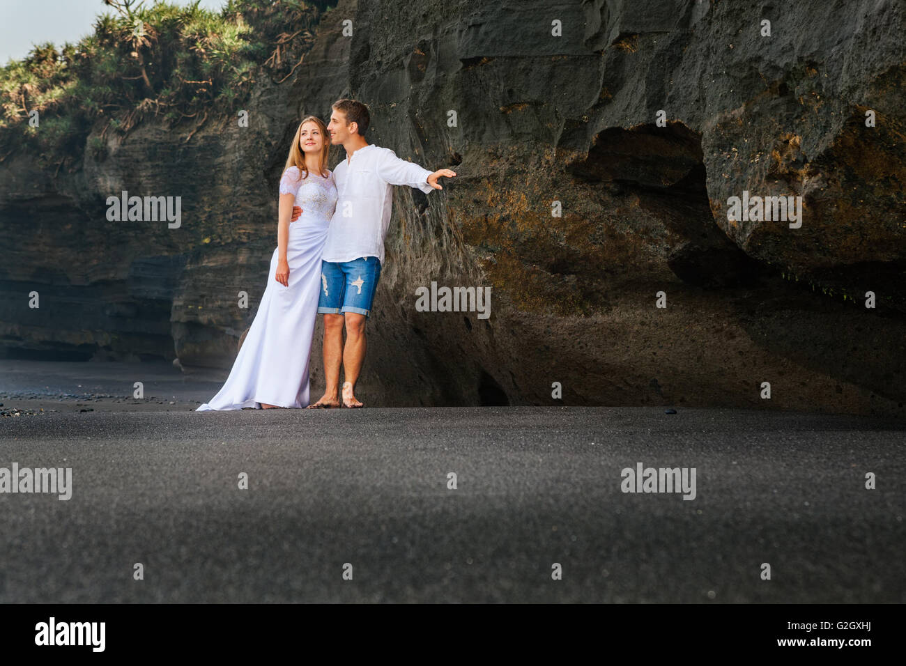 Felice famiglia giovane sulla luna di miele vacanze - appena sposato amare l uomo e la donna che abbraccia sulla spiaggia di sabbia con black rock sullo sfondo Foto Stock