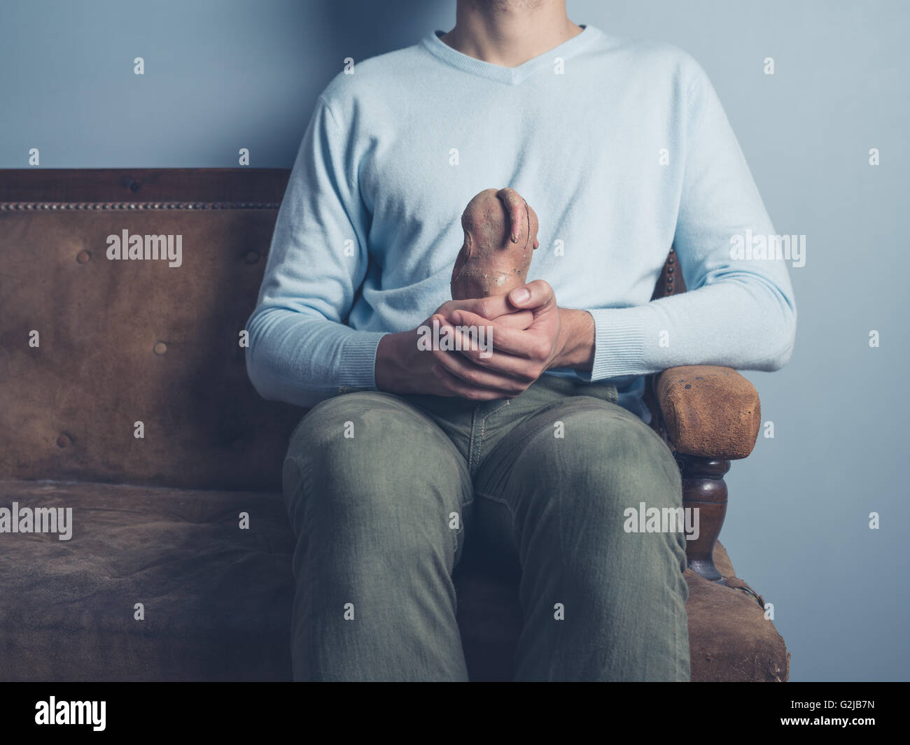 Un giovane uomo è seduto su un vecchio divano ed è in possesso di una strana patata dolce Foto Stock