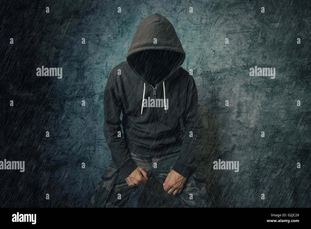 Spooky male persona criminale con giacca con cappuccio in piedi nella pioggia nella parte anteriore della parete di cemento. Foto Stock