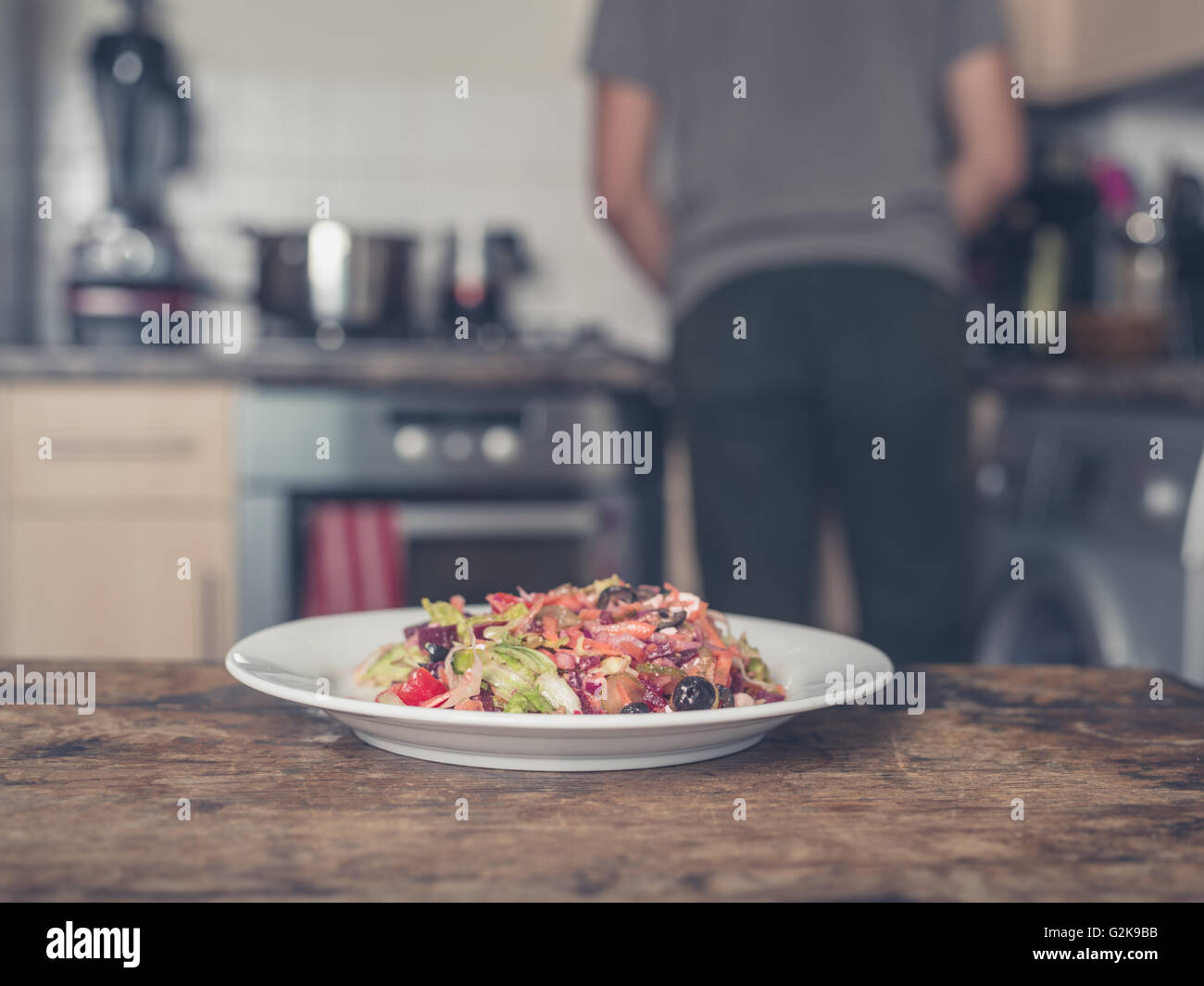 Una piastra con insalata su di una tavola in una cucina con un uomo per la cottura in background Foto Stock