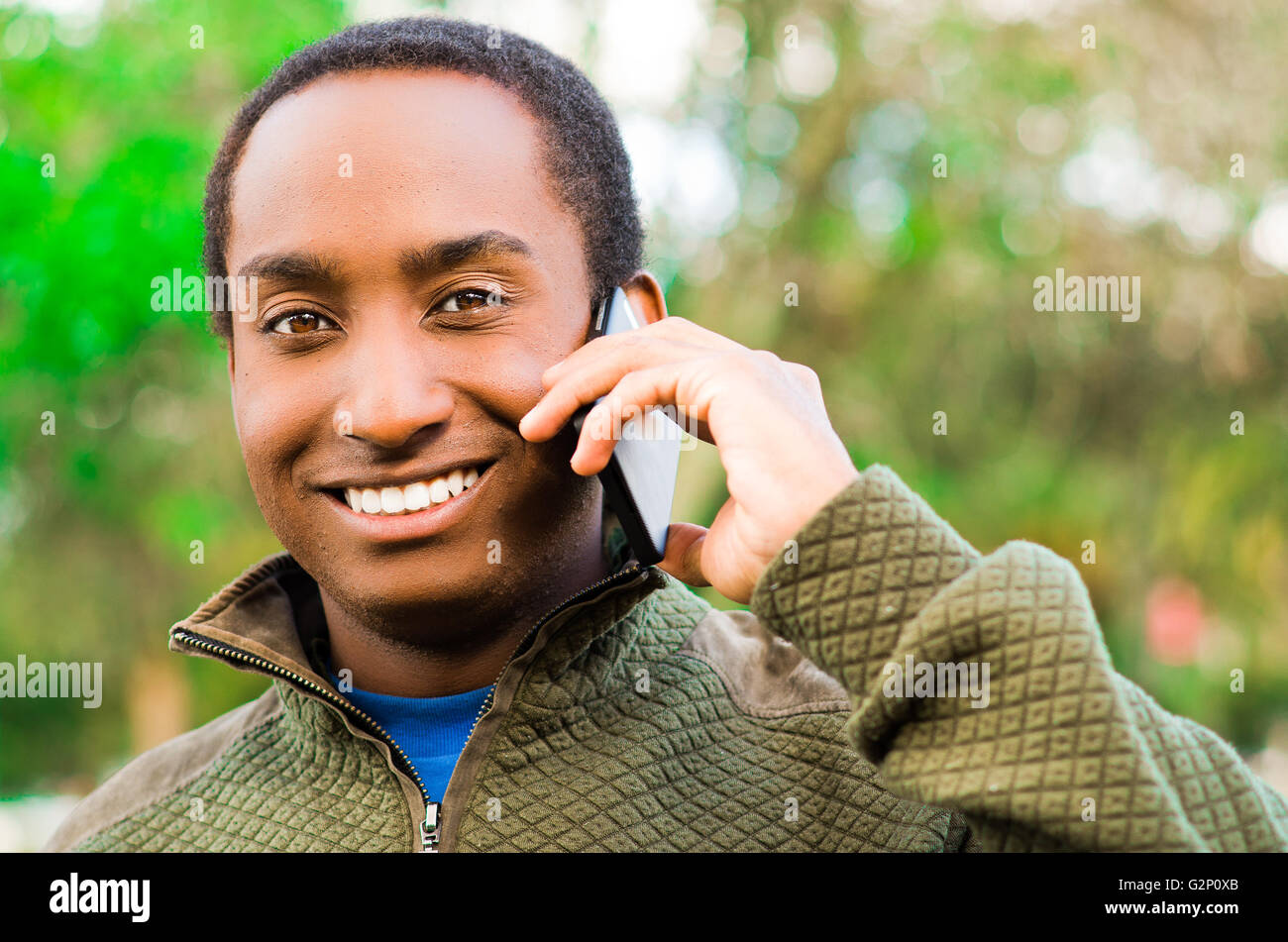 Bello ispanico uomo nero che indossa un maglione verde in all'aperto area parco tenendo in mano il telefono all'orecchio e parlare allegramente mentre ridono Foto Stock