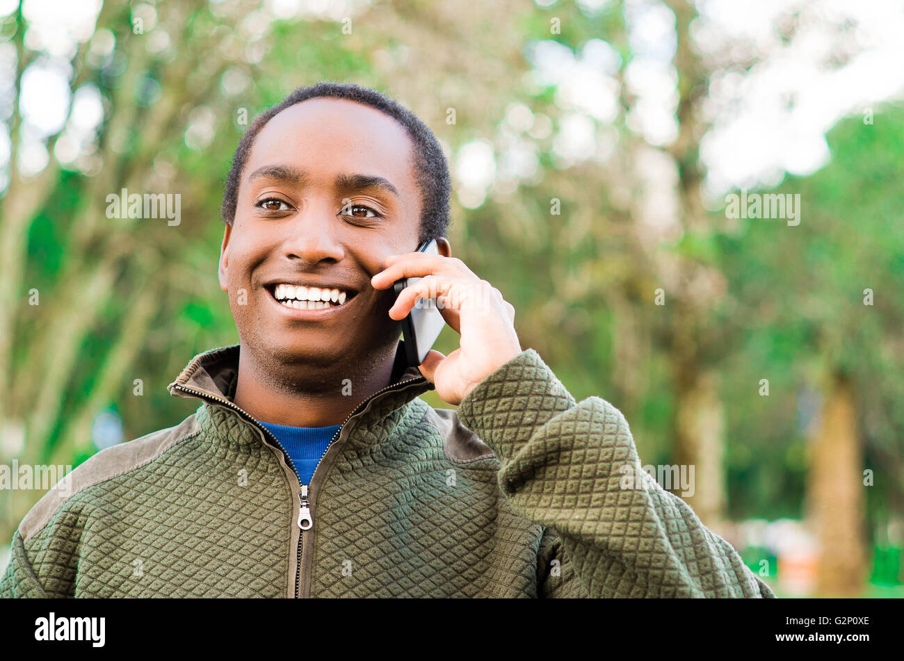 Bello ispanico uomo nero che indossa un maglione verde in all'aperto area parco tenendo in mano il telefono all'orecchio e parlare allegramente mentre ridono Foto Stock