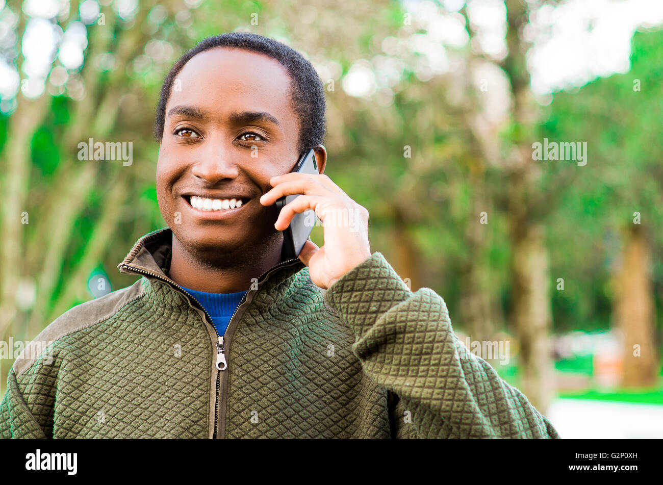 Bello ispanico uomo nero che indossa un maglione verde in all'aperto area parco tenendo in mano il telefono all'orecchio e parlare allegramente mentre ridono Foto Stock