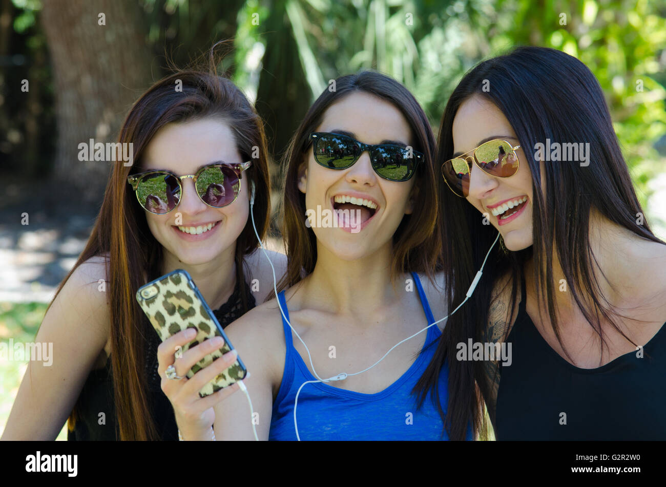 Tre giovani ragazze che consente di ascoltare musica sul proprio telefono sorridente in un parco in occhiali da sole Foto Stock