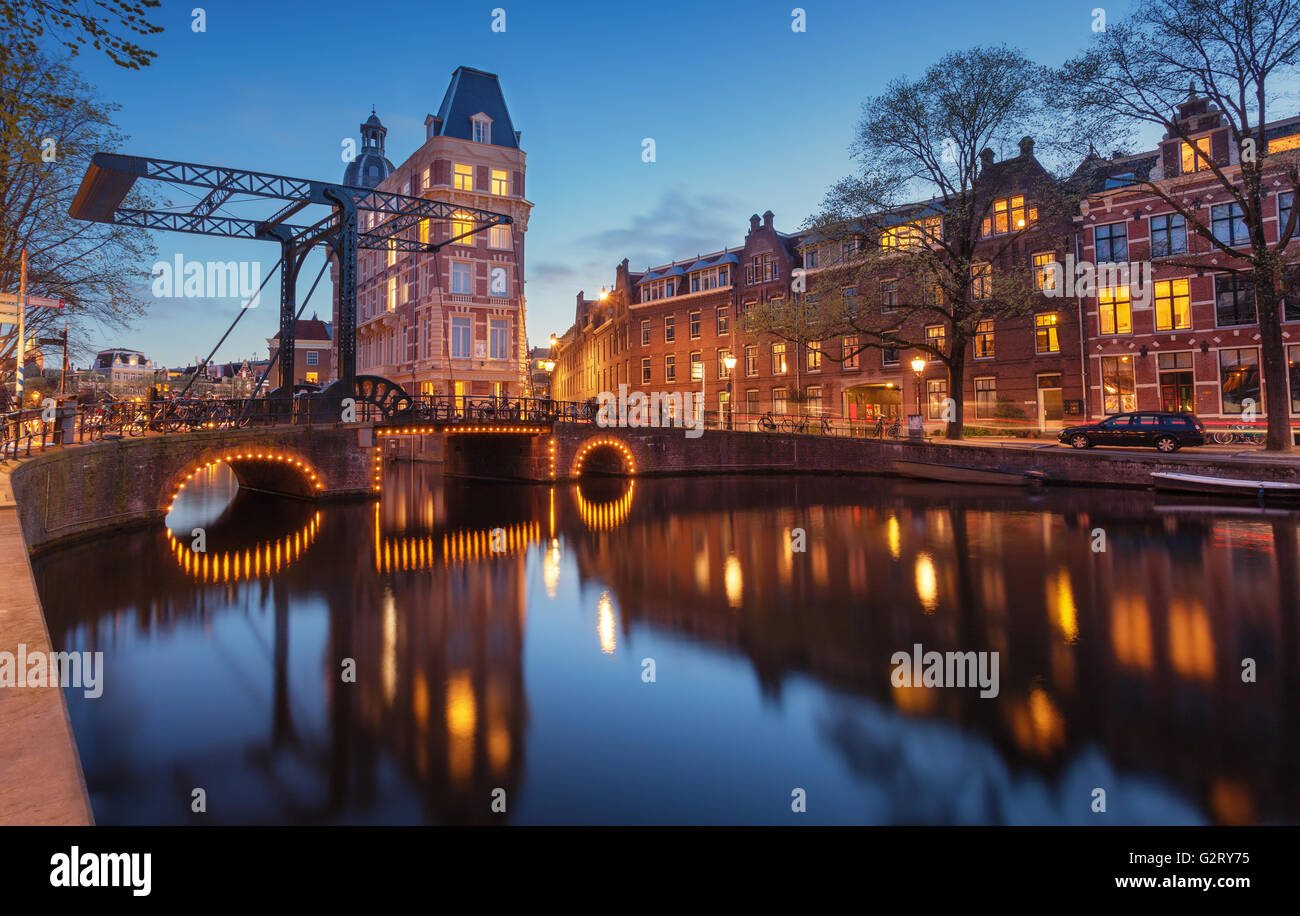 Bellissimo paesaggio urbano di notte a Amsterdam, Paesi Bassi. Riflette le luci della città in acqua con arco e cielo blu. Illuminazione notturna o Foto Stock