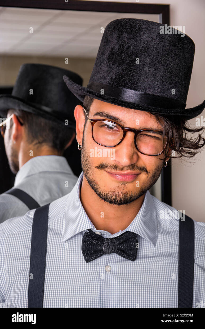 Ritratto di bruna giovane uomo in bicchieri, cappello, il filtro bow tie, calze autoreggenti e shirt guardando la fotocamera. Specchio dietro di lui Foto Stock