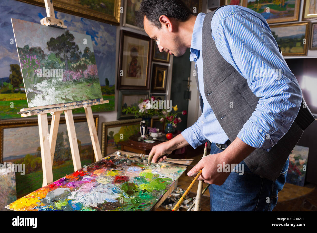 Artista professionale lavorando in una galleria o studio la selezione di una particolare tonalità di colore da una tavolozza colorata di miscele di olio vernici come egli lavora su un dipinto su un cavalletto. Foto Stock