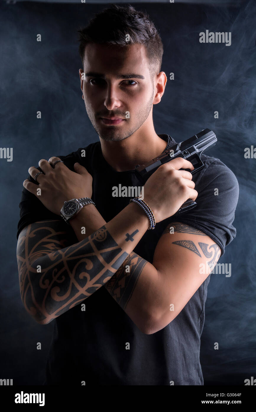 Giovane uomo bello tenendo una mano pistola, vestita di nero t-shirt, con le braccia incrociate sul petto e su sfondo scuro in studio Foto Stock