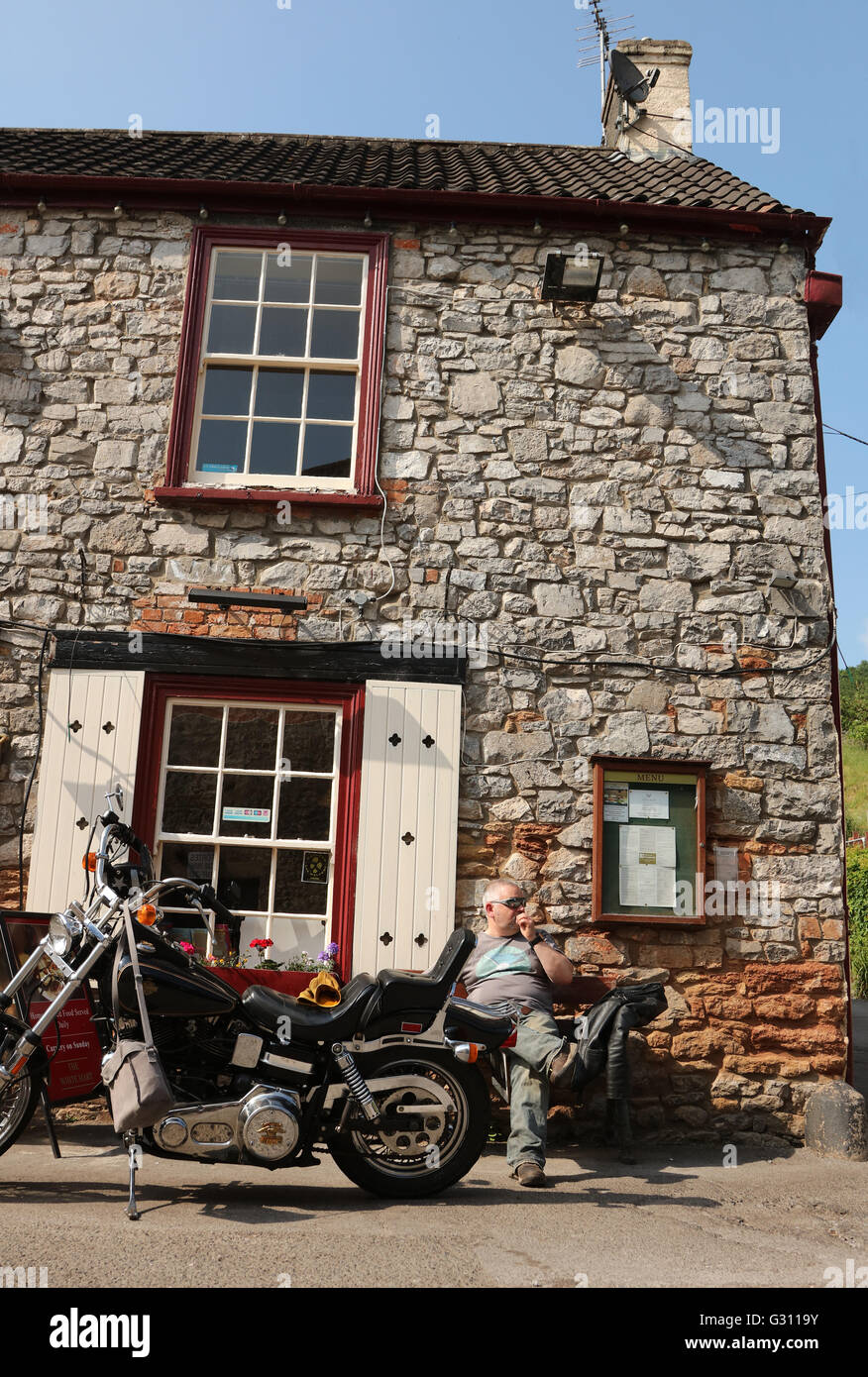 Giugno 2016 - ragazzo maturo e il suo grande Harley Davidson moto prendere una pausa al sole da un tradizionale Pub inglese di campagna Foto Stock