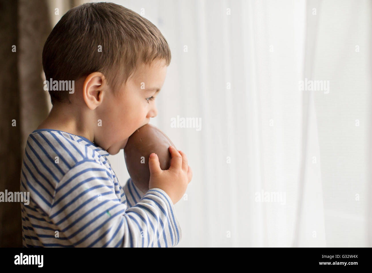 Ragazzo in piedi dalla finestra di mangiare una grande cioccolato uovo di pasqua Foto Stock