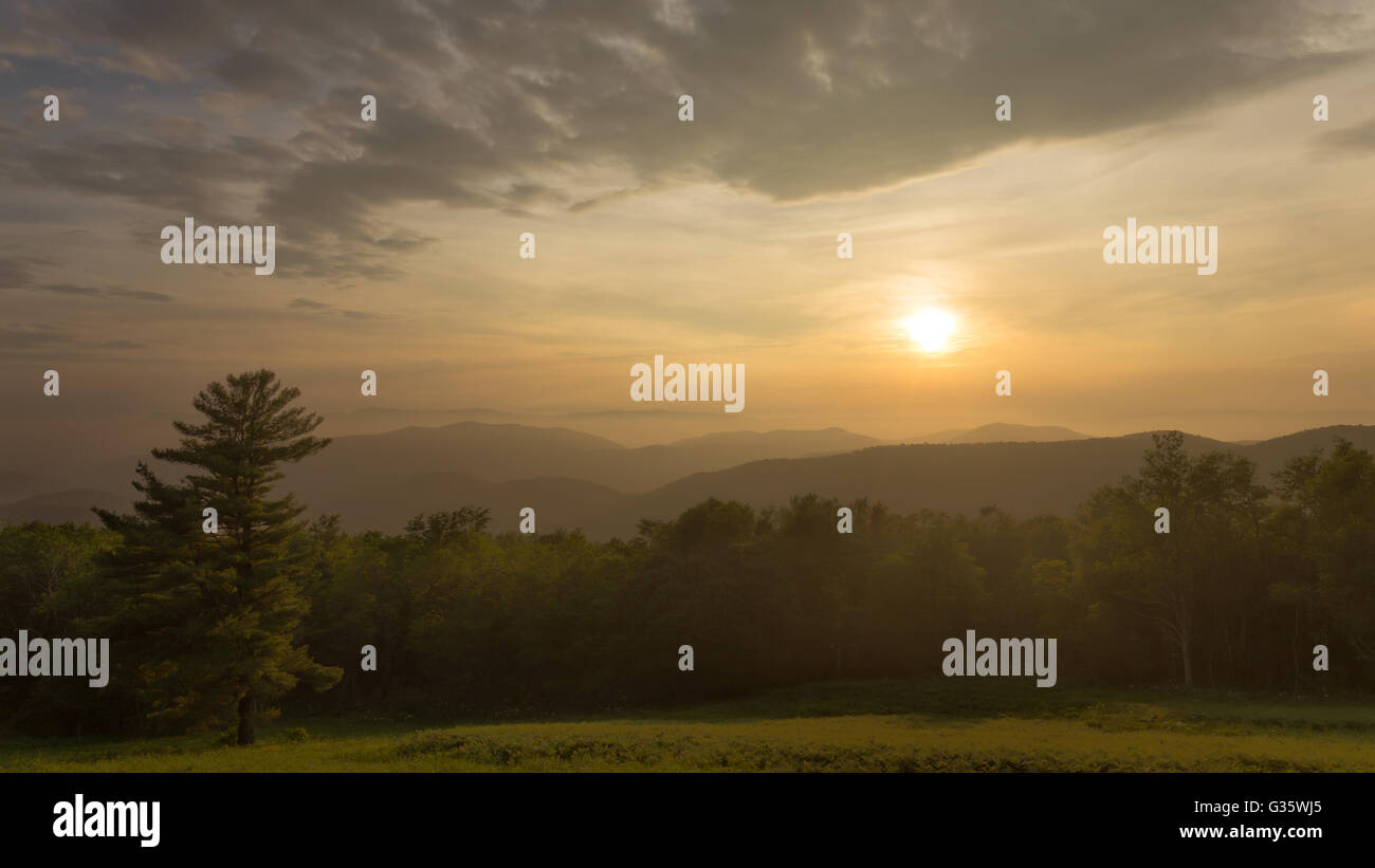 Il sole tramonta oltre le Blue Ridge Mountains nel Parco Nazionale di Shenandoah come le nuvole creano una luce calda sopra il prato Foto Stock
