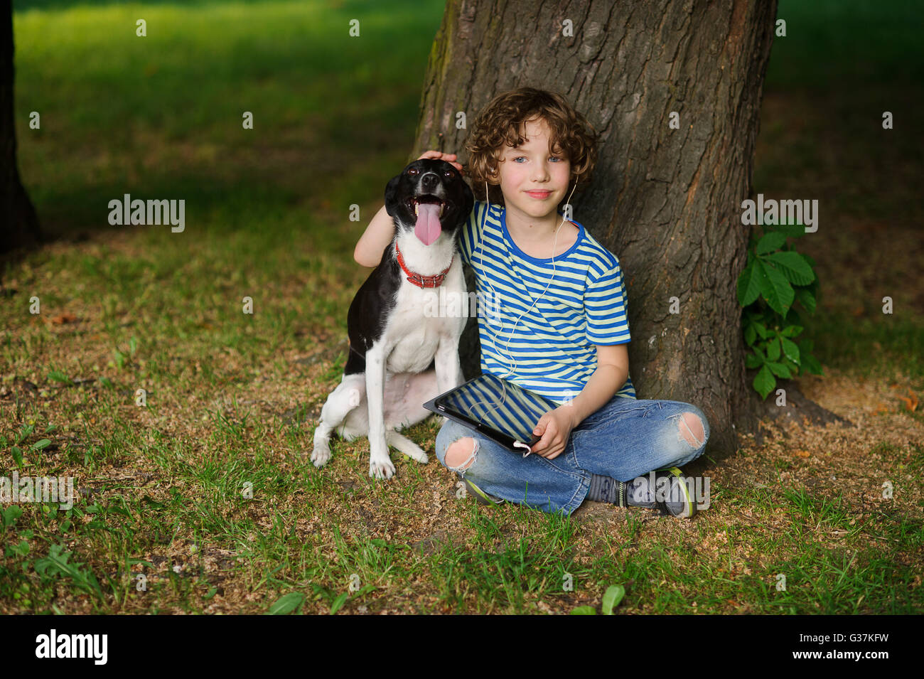 Il ragazzo con un cane si siede sulla terra sotto un albero. Il bambino si siede con le gambe incrociate. Egli ha messo una mano sulla testa di un animale domestico. 0 Foto Stock