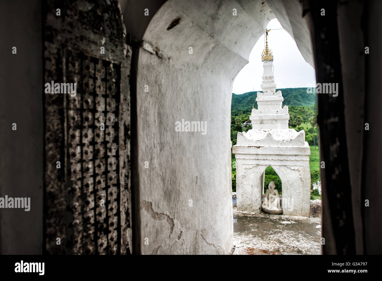 Statua della Pagoda di Hsinbyume Mingun Myanmar // MINGUN, Myanmar — Una vista attraverso una porta rivela l'hti e una piccola struttura contenente una statua nella Pagoda di Hsinbyume. Costruita nel 1816, la pagoda si trova a Mingun vicino a Mandalay ed è stata progettata come una rappresentazione del Monte Meru, la montagna sacra della cosmologia buddista. La caratteristica struttura dipinta di bianco presenta sette livelli terrazzati con forme ondulate che la distinguono dalla tradizionale architettura a pagoda. La Pagoda di Hsinbyume fu commissionata dal re Bagyidaw come memoriale della sua prima moglie, la principessa Hsinbyume, che morì a Chil Foto Stock
