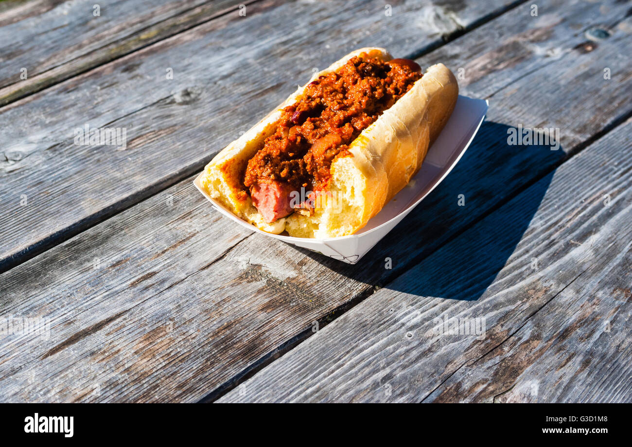 In parte mangiato salsiccia coperta in hot chili su un panino in un vassoio della carta, su weathered grigio Tavolo picnic. Foto Stock