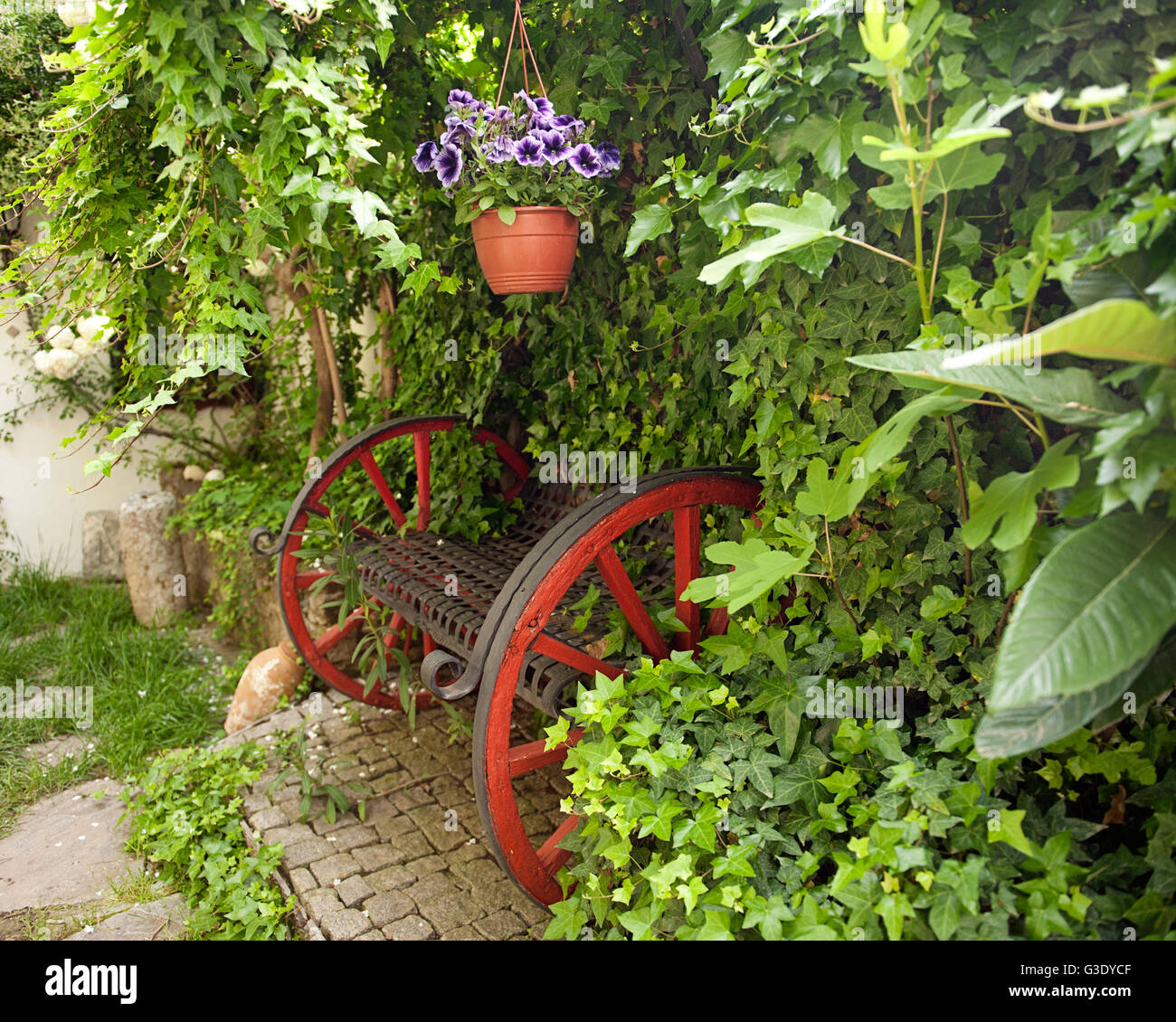 Una panchina da giardino rustica realizzata in un carrello di legno si trova sotto l'edera verde con un vaso di fiori appeso, creando un tranquillo spazio all'aperto. Foto Stock