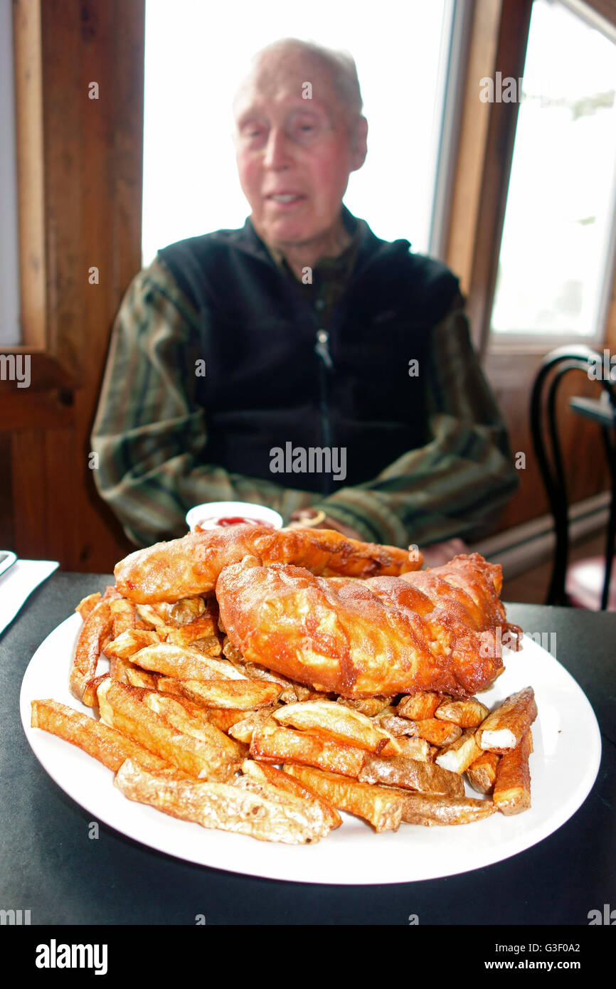 Un anziano uomo seduto dietro a un piatto di pesce e patatine fritte su un tavolo Foto Stock