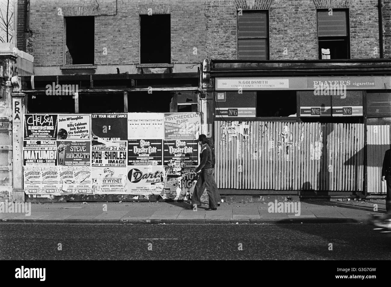 Immagine di archivio di un uomo in flares e una donna passato a piedi negozi abbandonati intonacate con manifesti pubblicitari, Lambeth, Londra, Inghilterra, 1979 Londra 1970s Foto Stock