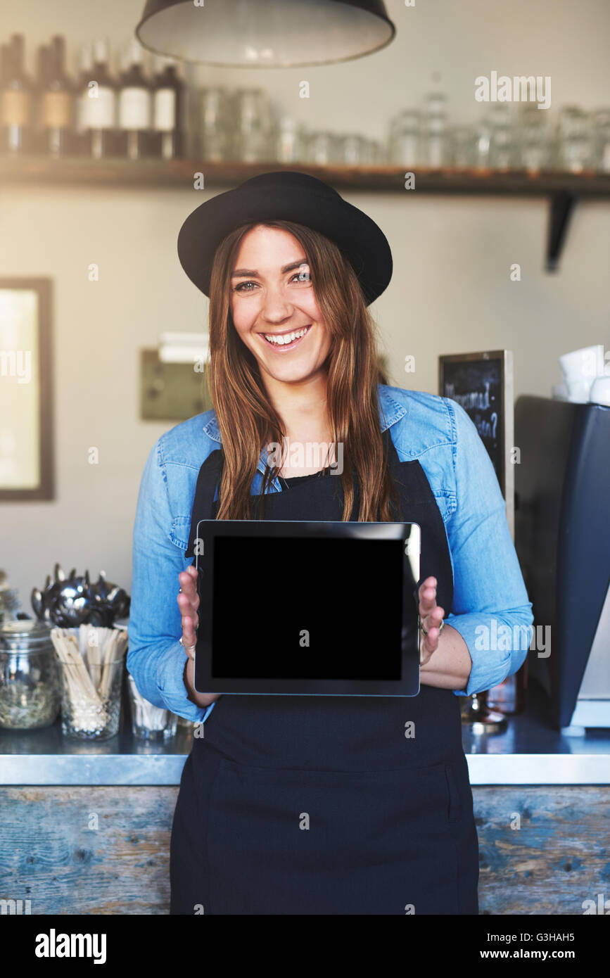 Imprenditore femmina indossando cappello nero e grembiule sorrisi a telecamera tenendo il computer tablet verso l'esterno fra entrambe le mani Foto Stock