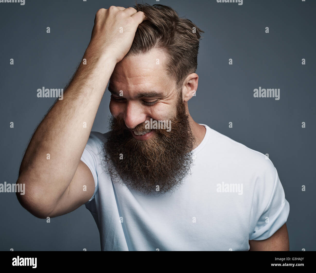Ridendo giovane uomo con la barba lunga e la camicia bianca tenendo il pelo mentre è rivolto verso il basso su uno sfondo grigio Foto Stock
