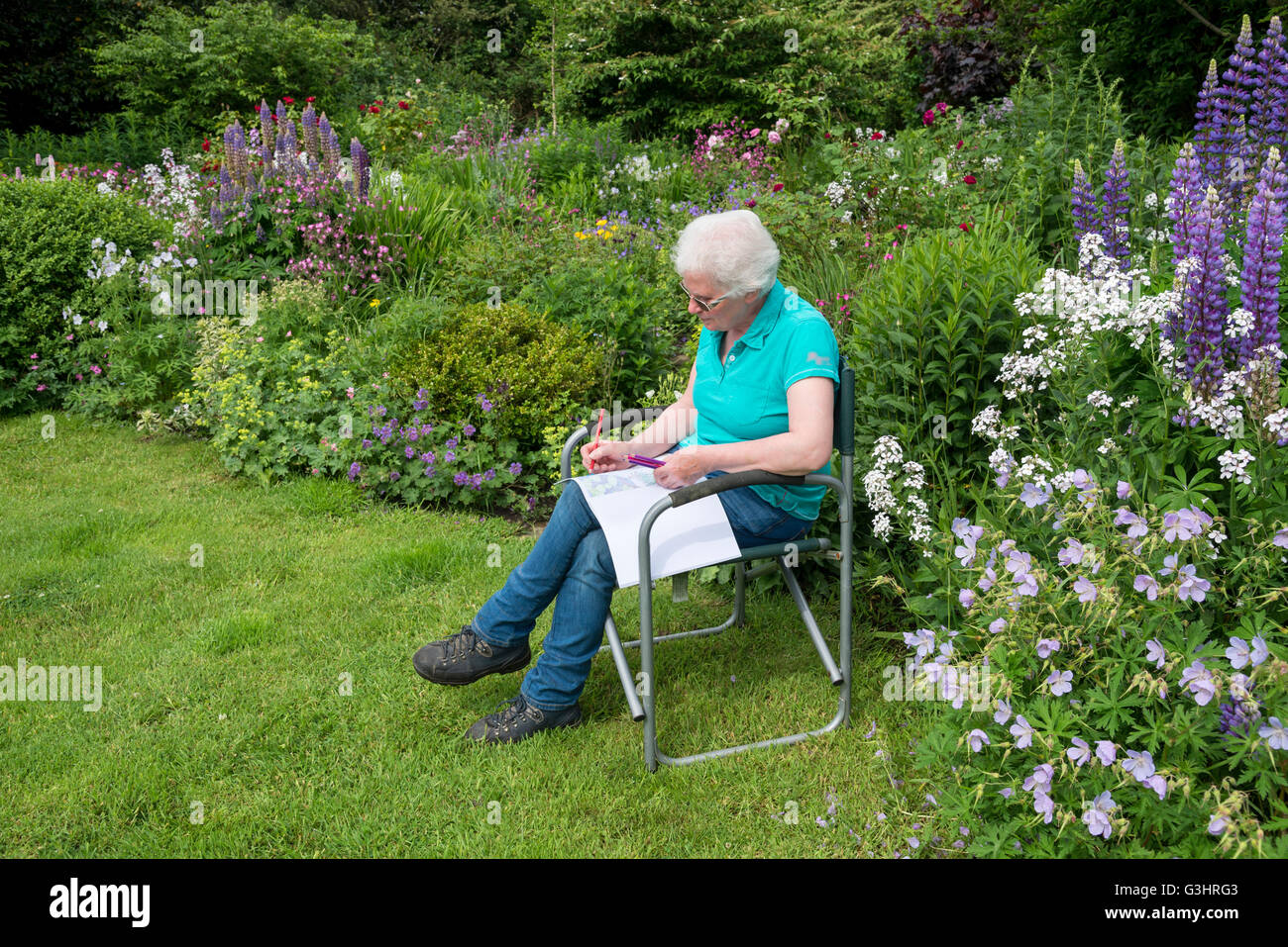 Una donna matura si rilassa in un giardino di campagna con un adulto libro da colorare circondato da fiori d'estate. Foto Stock