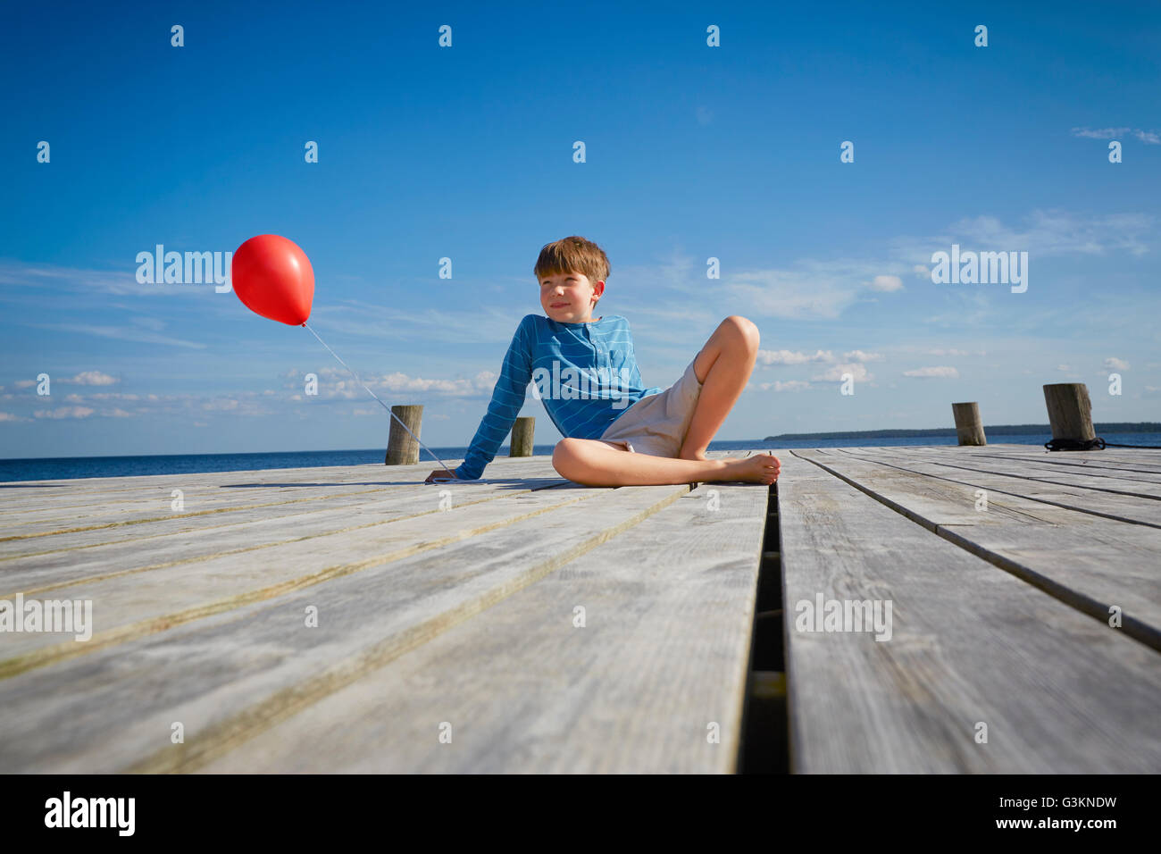 Ragazzo seduto sul molo in legno, tenendo rosso palloncino elio Foto Stock