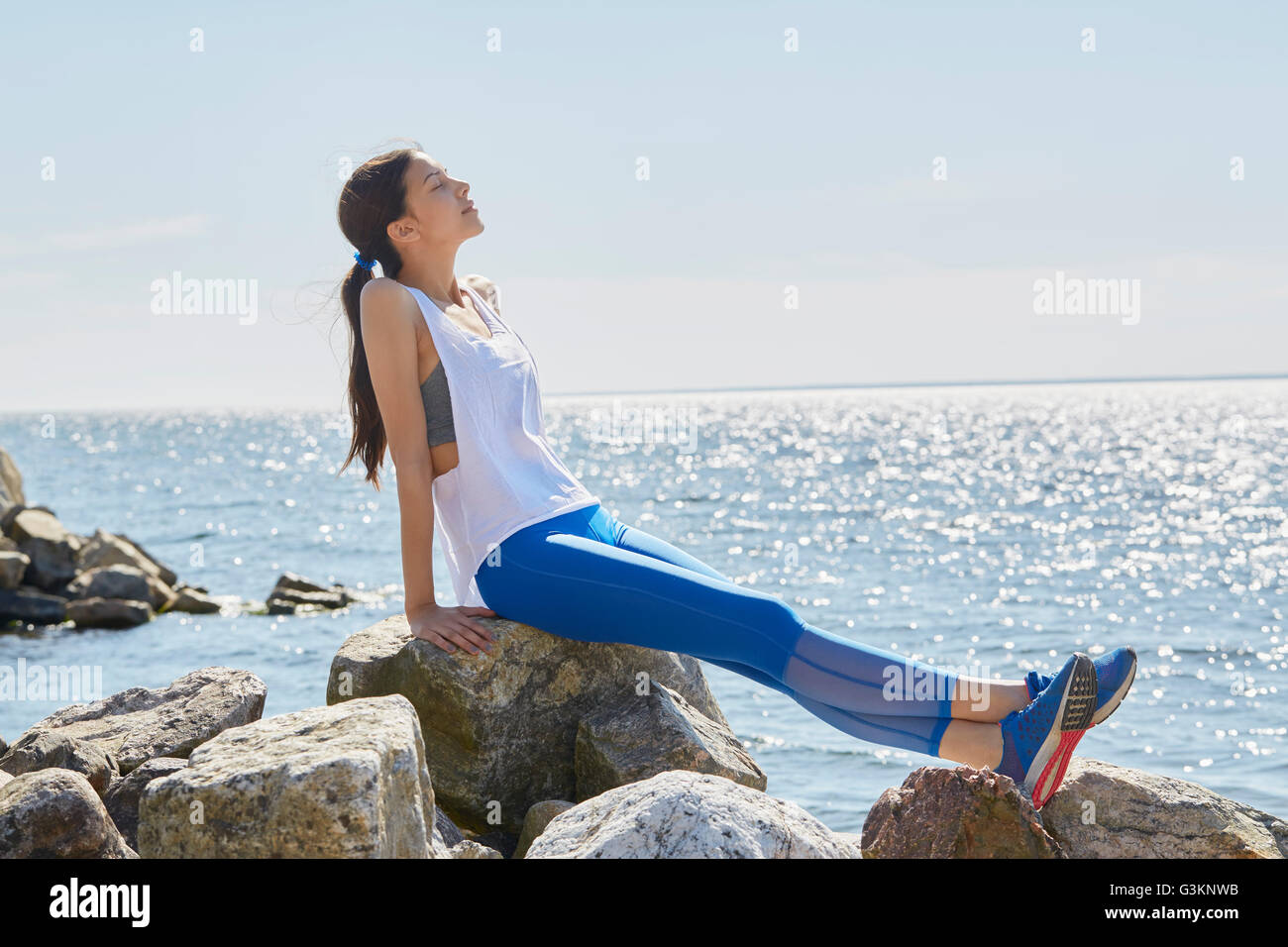 Vista laterale della donna che indossa abbigliamento sportivo seduti sulle rocce crogiolarsi nella luce solare Foto Stock