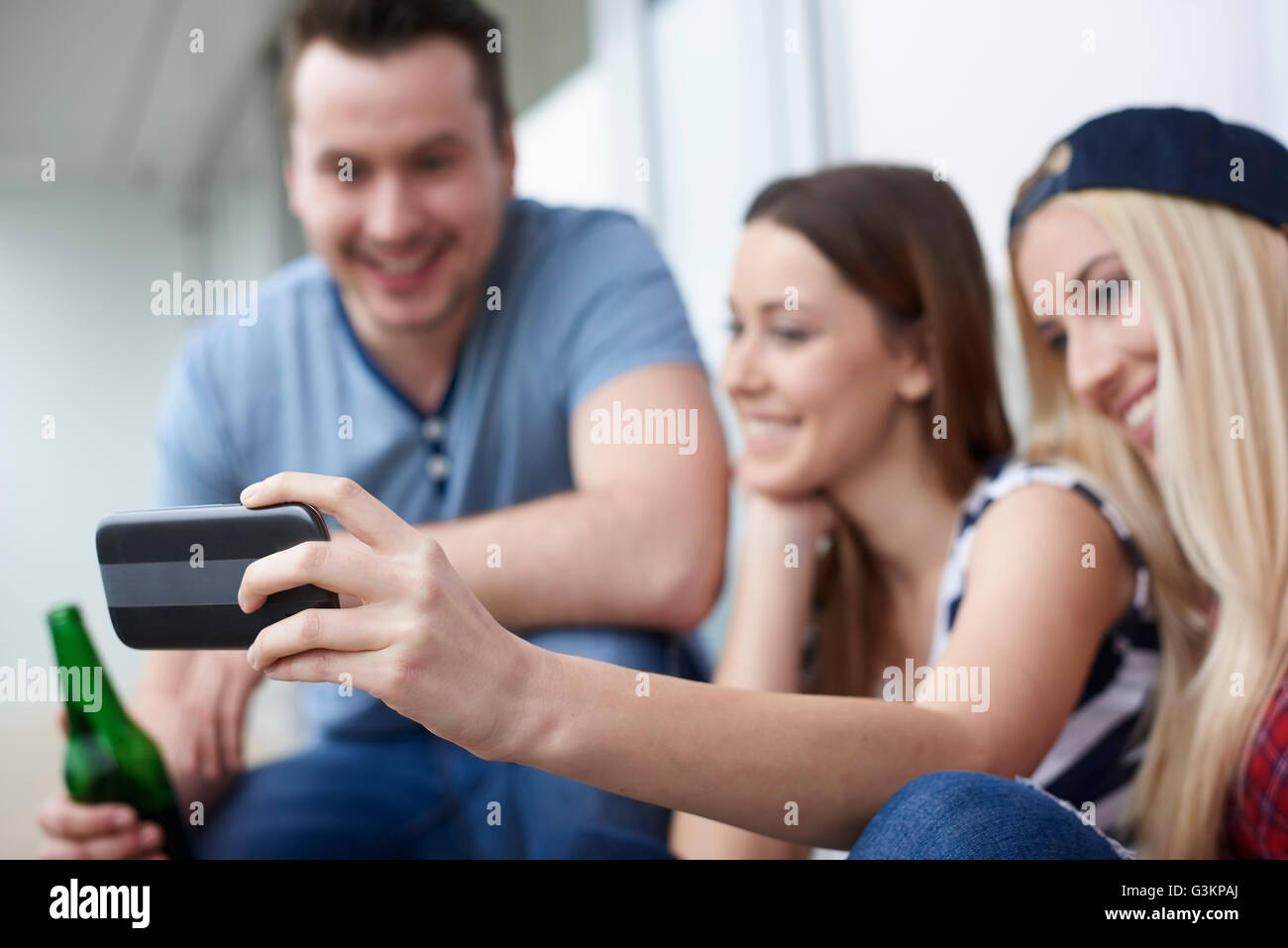 Gruppo di amici seduti all'aperto e di bere la birra in bottiglia, tenendo autoritratto, utilizza lo smartphone Foto Stock