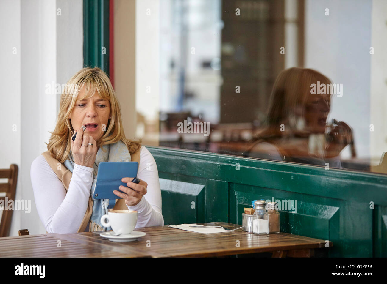 Donna matura utilizza lo smartphone per applicare il rossetto al cafè sul marciapiede tabella Foto Stock