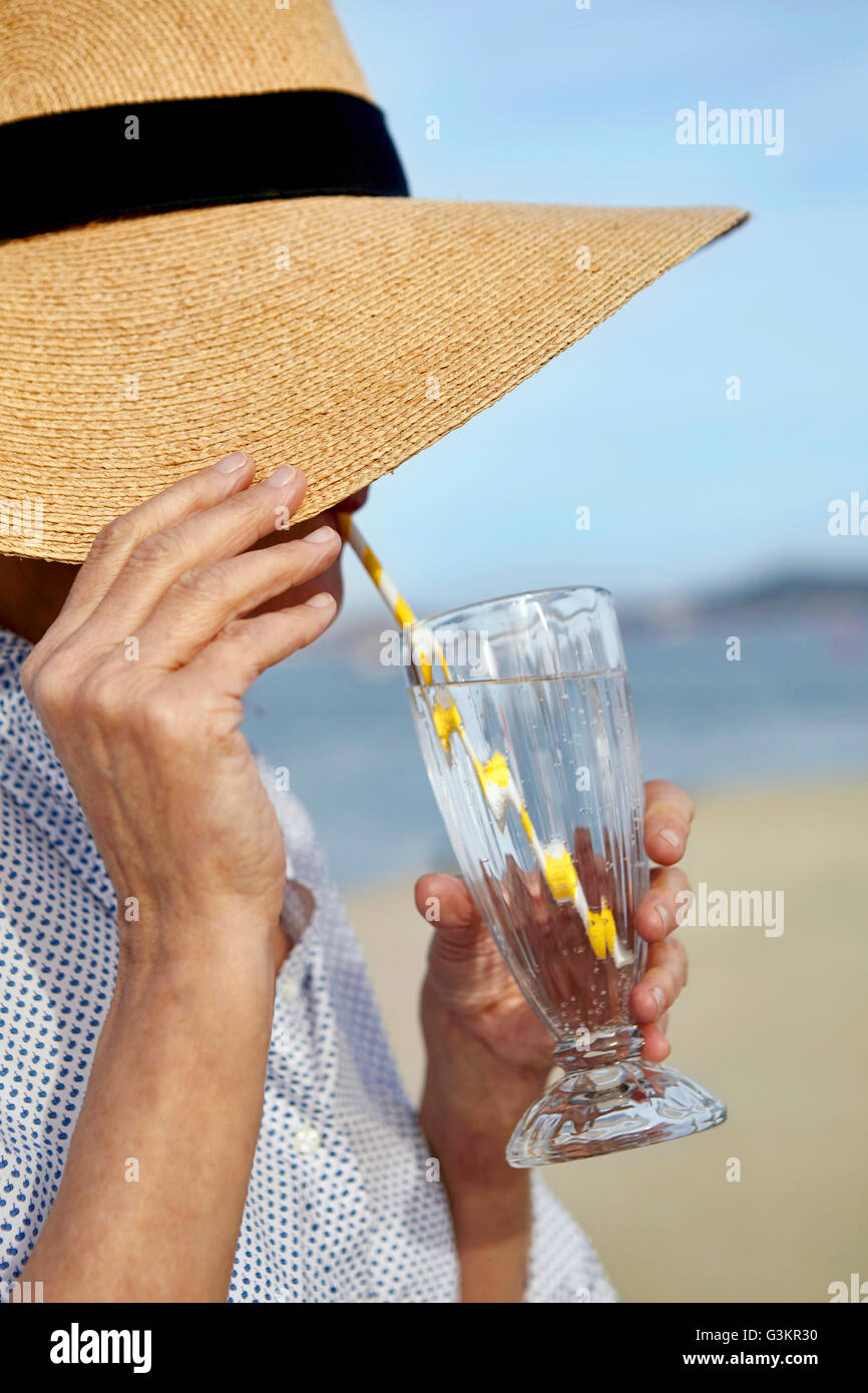 Donna matura indossando cappello di paglia, accanto alla spiaggia, bere drink fresco attraverso la cannuccia per bevande Foto Stock