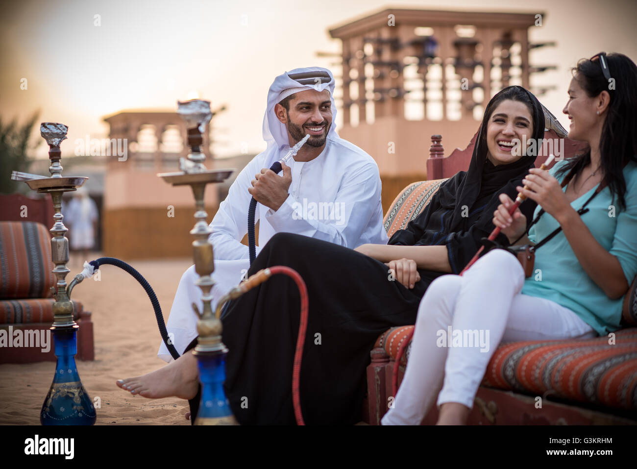 Locale giovane indossando abiti tradizionali di fumare shisha sul divano con turista femminile, Dubai, Emirati Arabi Uniti Foto Stock